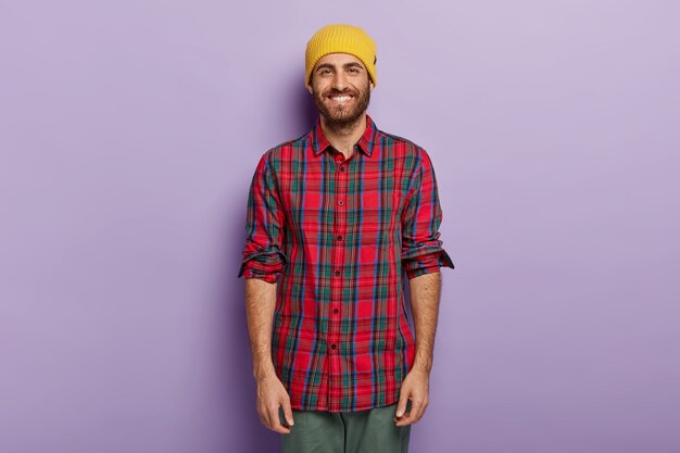 Cara feliz com um sorriso brilhante, barba por fazer, usa chapéu amarelo e camisa xadrez, aproveita o tempo livre