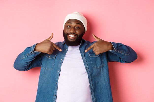 Cara feliz jovem hipster afro-americana apontando para a camiseta de si mesmo, sorrindo satisfeito, em pé sobre um fundo rosa.