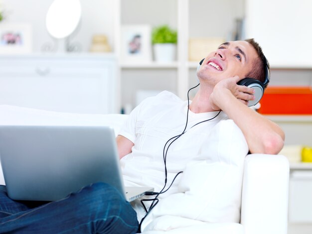 Cara feliz ouvindo música no fone de ouvido do laptop e deitado no sofá