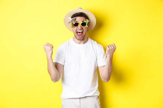 Cara feliz saindo de férias, vencendo ou comemorando, usando óculos escuros e chapéu de verão. Turista parece animado, de pé contra um fundo amarelo.