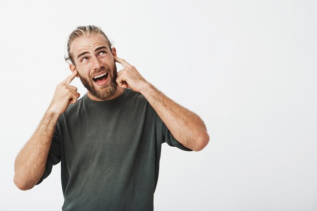 Cara jovem bonita com corte de cabelo elegante e barba, conectando as orelhas com os dedos