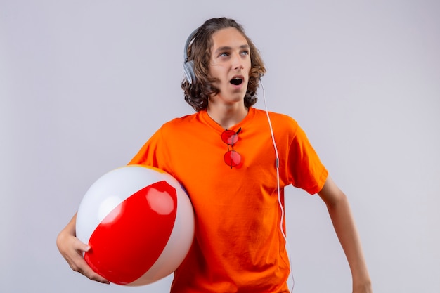 Foto grátis cara jovem bonito de camiseta laranja com fones de ouvido segurando uma bola inflável, olhando de lado em pé surpreso e espantado
