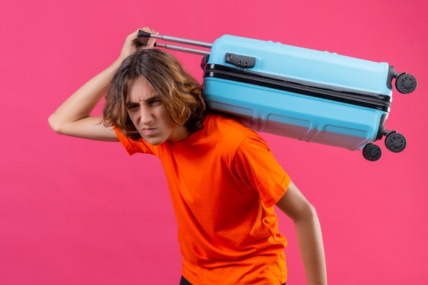 Foto grátis cara jovem bonito de camiseta laranja segurando a mala de viagem, olhando em pé entediado e cansado