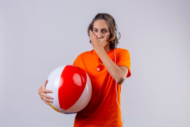 Cara jovem bonito de camiseta laranja, segurando uma bola inflável com fones de ouvido espantado e surpreso cobrindo a boca com a mão em pé