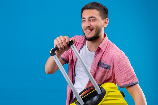 Cara jovem bonito viajante segurando a mala, olhando de lado positivo e feliz sorrindo em pé sobre fundo azul