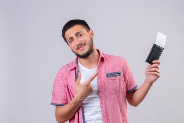 Cara jovem bonito viajante segurando passagens aéreas, apontando com o dedo para eles, olhando para a câmera com sorriso confiante no rosto feliz e positivo em pé sobre fundo branco