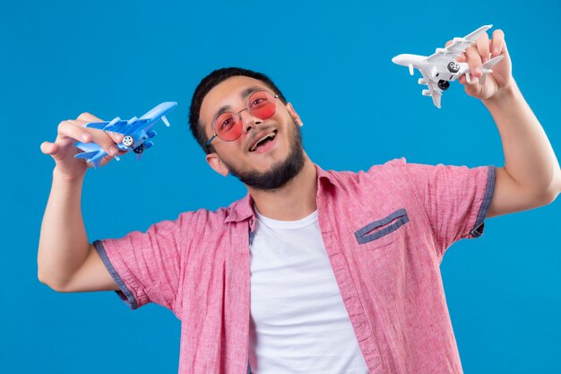 Cara jovem bonito viajante usando óculos escuros segurando aviões de brinquedo brincando com eles, olhando feliz e positivo, sorrindo alegremente em pé sobre fundo azul