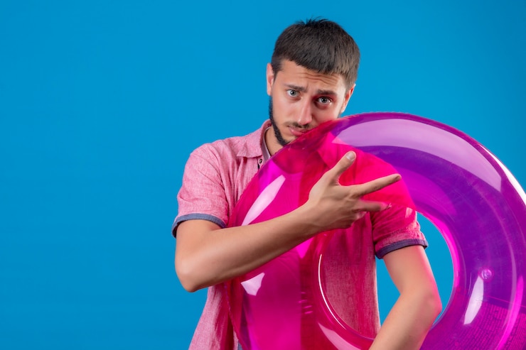Foto grátis cara jovem bonito viajante usando óculos escuros, segurando o anel inflável, olhando para a câmera com expressão triste em pé sobre fundo azul