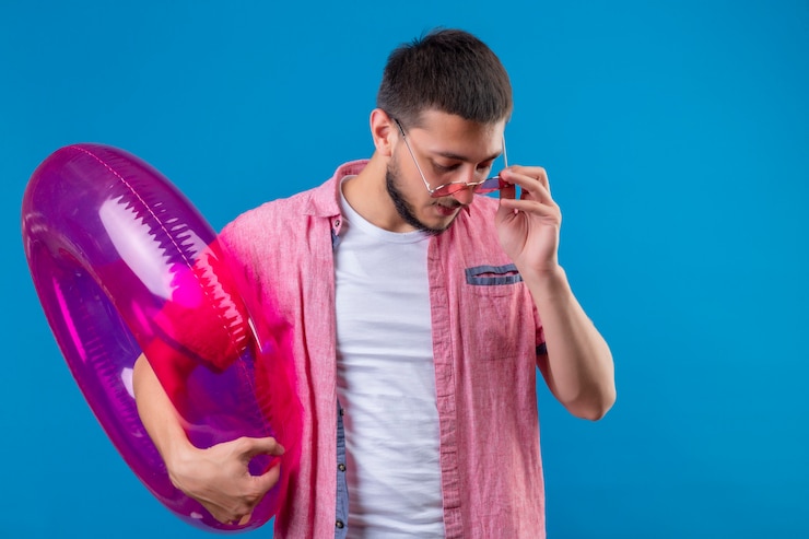 Foto grátis cara jovem bonito viajante usando óculos escuros, segurando o anel inflável, olhando para baixo com expressão séria confiante em pé sobre fundo azul