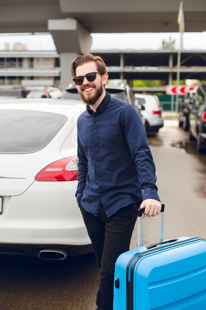 Cara jovem com barba em óculos de sol pretos está de pé com a mala na zona de estacionamento no aeroporto. Ele veste camisa preta com calça e sorri para a câmera.