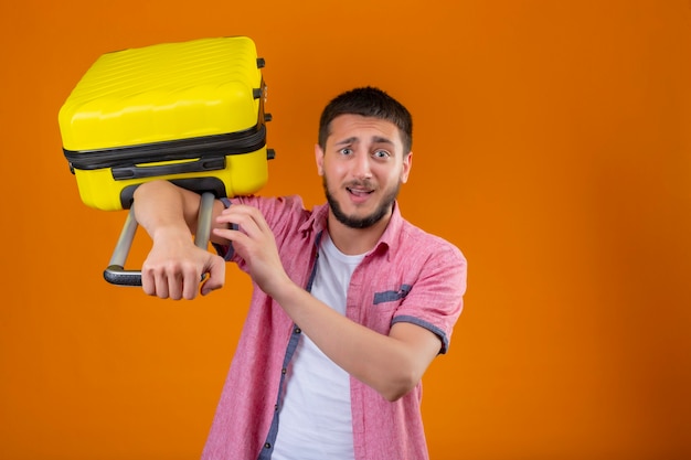 Foto grátis cara jovem e bonito viajante chateado segurando uma mala, olhando para a câmera com uma expressão triste em pé sobre um fundo laranja