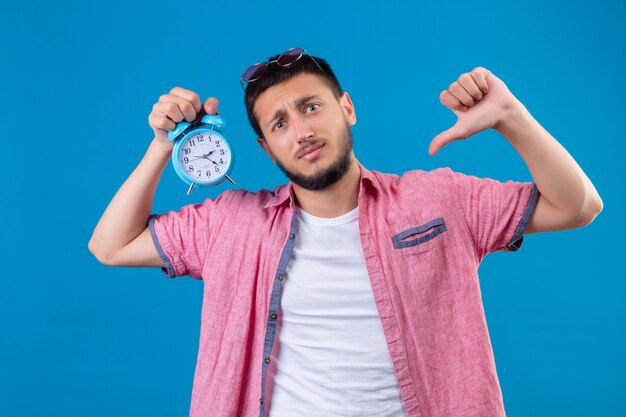 Foto grátis cara jovem e bonito viajante segurando um despertador, olhando para a câmera com uma expressão triste no rosto, mostrando os polegares para baixo em pé sobre um fundo azul