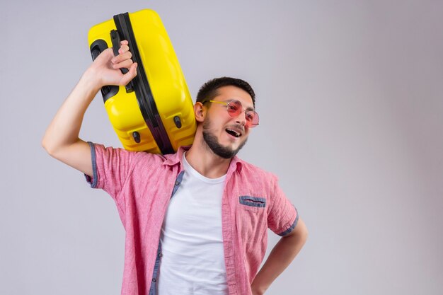 Cara jovem e bonito viajante segurando uma mala, olhando de lado, sorrindo, positivo e feliz em pé sobre um fundo branco