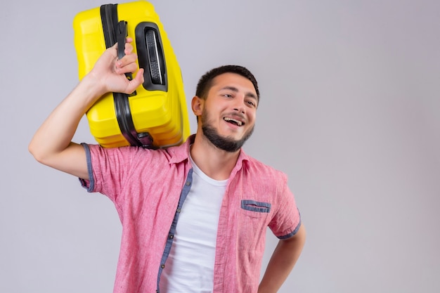 Cara jovem e bonito viajante segurando uma mala, olhando de lado, sorrindo, positivo e feliz em pé sobre um fundo branco