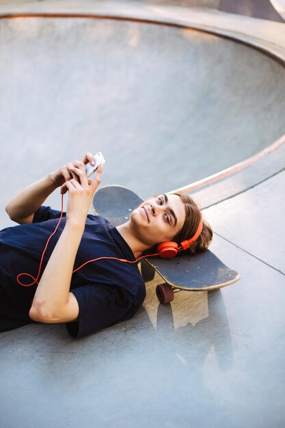 Cara jovem em fones de ouvido laranja deitado no skate com celular nas mãos enquanto alegremente olhando na câmera no skatepark isolado