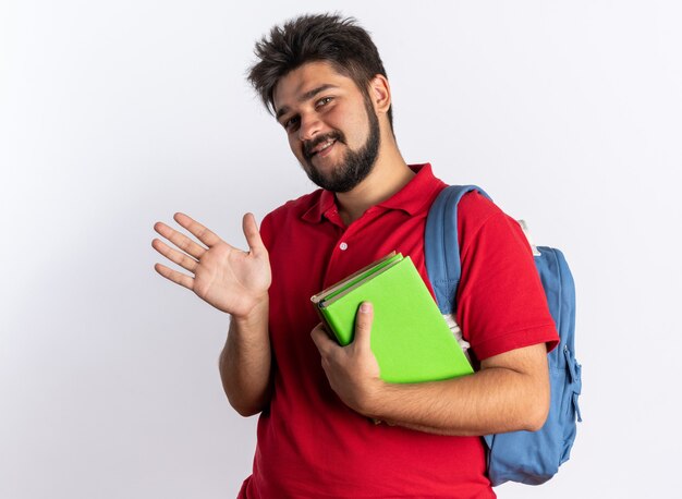 Cara jovem estudante barbudo em uma camisa pólo vermelha com mochila segurando notebooks feliz e alegre sorrindo acenando com a mão em pé sobre uma parede branca