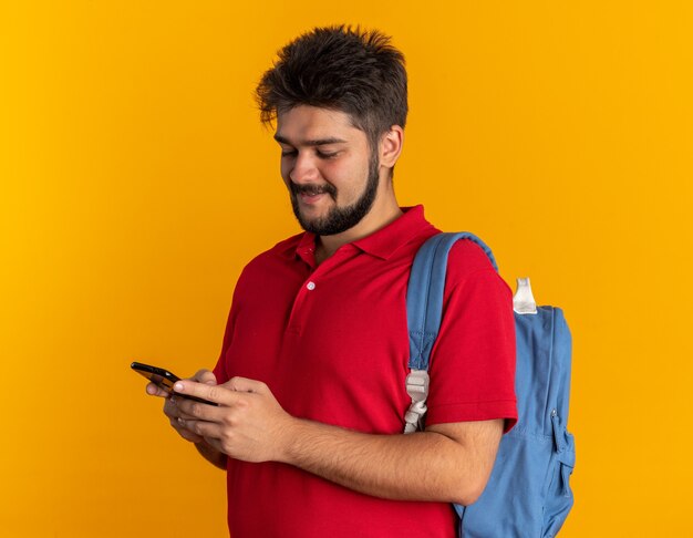 Cara jovem estudante barbudo em uma camisa pólo vermelha com mochila segurando um smartphone olhando para ele sorrindo alegremente feliz e positivo em pé sobre a parede laranja