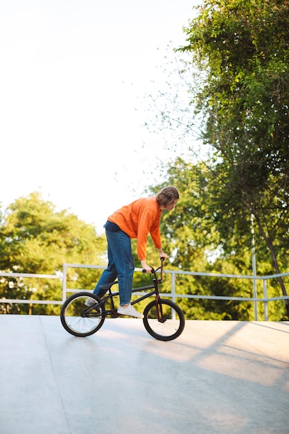 Foto grátis cara jovem legal no pulôver laranja andando de bicicleta e praticando acrobacias no skatepark moderno