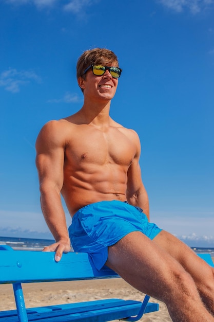 Foto grátis cara jovem musculoso incrível em shorts de natação azul, sentado no banco sobre o mar e o céu azul.