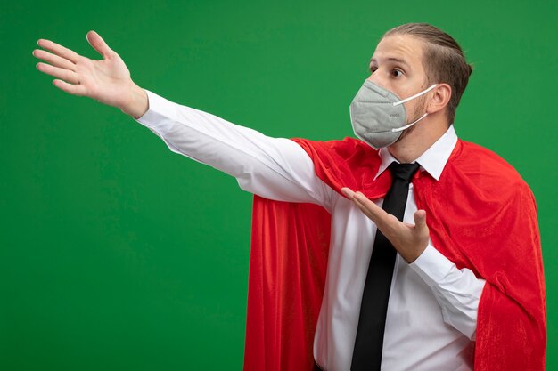 Foto grátis cara jovem super-herói surpreso usando máscara médica, olhando para o lado e pontos com as mãos ao lado isoladas sobre fundo verde