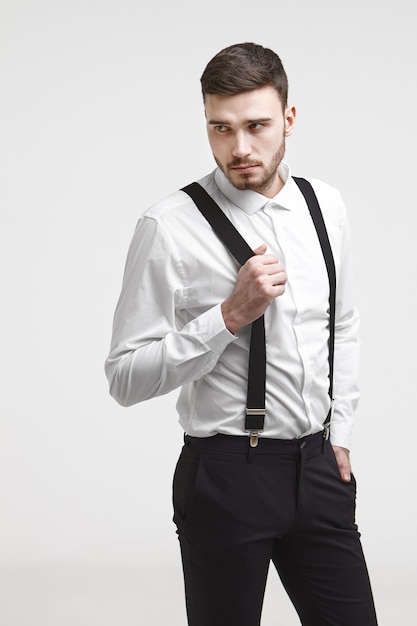 Foto grátis cara na moda hippie com barba por fazer e corte de cabelo elegante, posando na parede do estúdio em branco, desviando o olhar com uma expressão misteriosa. jovem bonito barbudo do sexo masculino vestido com roupa formal puxando suspensórios