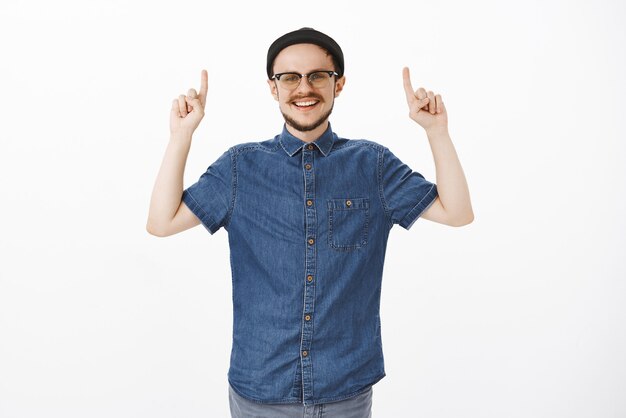 Cara oferecendo espaço de cópia legal lugar em pé com um gorro preto moderno e óculos sorrindo alegremente levantando os braços e apontando para cima de bom humor enquanto posava sozinho