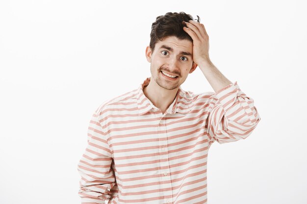 Cara pedindo desculpas, admitindo culpa. Retrato de modelo masculino atraente e inseguro e envergonhado, com barba e bigode, tocando o cabelo e sorrindo sem jeito, sendo esquecido ou tímido sobre parede cinza