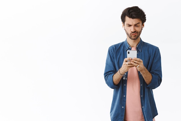 Cara perplexo tentando desculpas de maquiagem enquanto escrevia a mensagem segurando o sorriso do smartphone intrigado franzindo a testa olhando sério e determinado display de celular em pé fundo branco