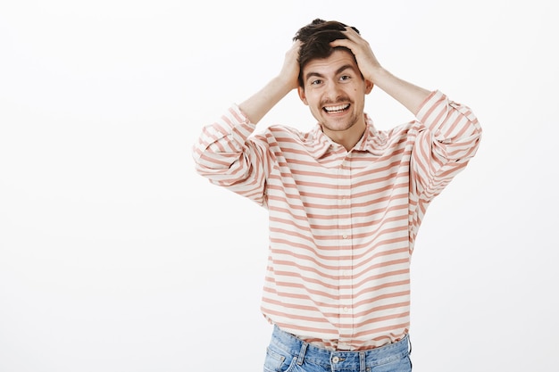 Foto grátis cara positivo e amigável nunca fica chateado facilmente. retrato de um homem bonito e feliz com barba e bigode, segurando as mãos na cabeça e sorrindo enquanto se lembra de algo que esqueceu sobre a parede cinza