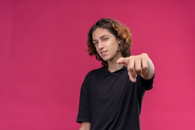 Cara sorridente com cabelo comprido e camiseta preta apontando para frente na parede rosa