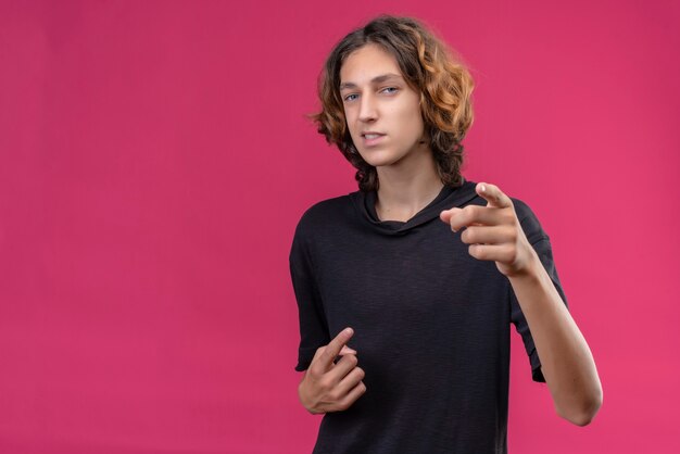 Cara sorridente com cabelo comprido e camiseta preta apontando para frente na parede rosa