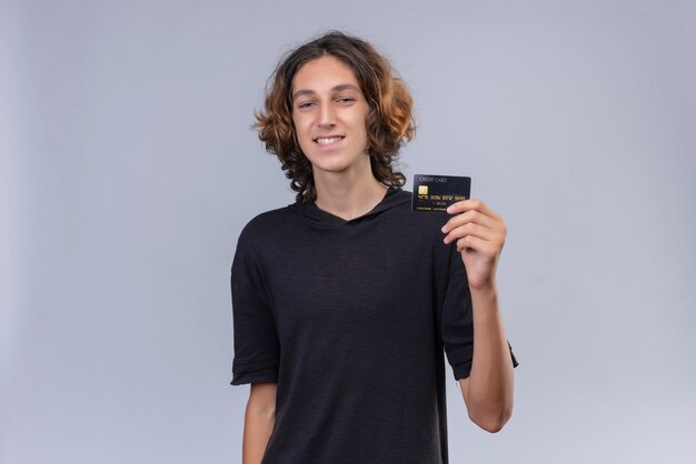 Cara sorridente com cabelo comprido e camiseta preta segurando um cartão do banco na parede branca