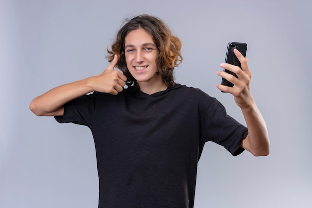 Cara sorridente com cabelo comprido e camiseta preta segurando um telefone e mostrando o polegar na parede branca