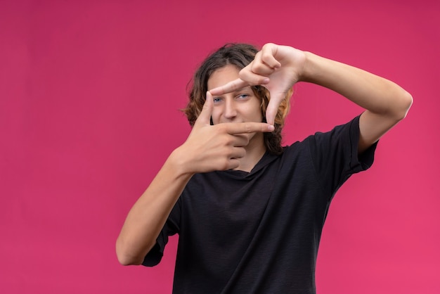 Cara sorridente com cabelo comprido e camiseta preta tirando uma foto na parede rosa