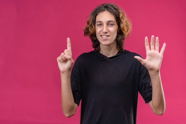 Cara sorridente com cabelo comprido em uma camiseta preta mostrando uma com uma mão e as outras cinco na parede rosa