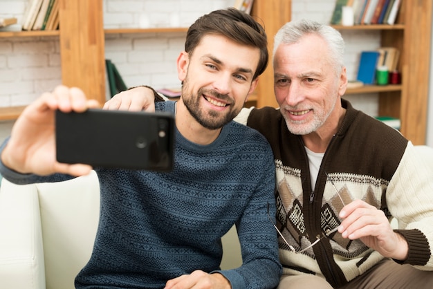 Foto grátis cara sorridente jovem e envelhecido homem alegre tomando selfie em smartphone no sofá