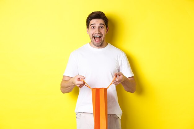 Cara surpreso abre a sacola de compras com o punho, parecendo animado e feliz para a câmera, em pé contra um fundo amarelo.