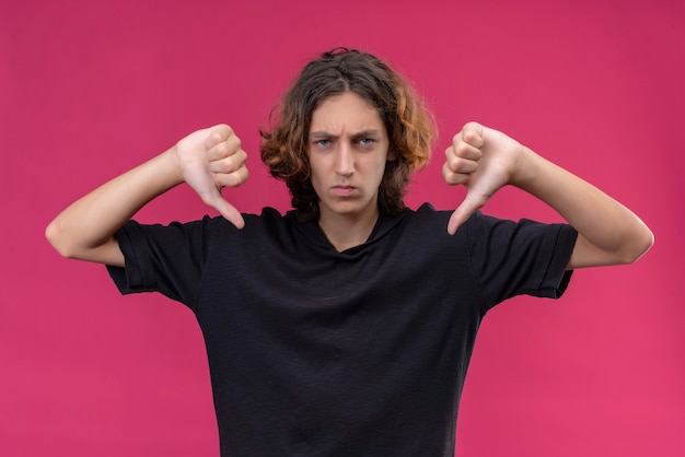 Cara zangado com cabelo comprido e camiseta preta mostrando o polegar para baixo na parede rosa