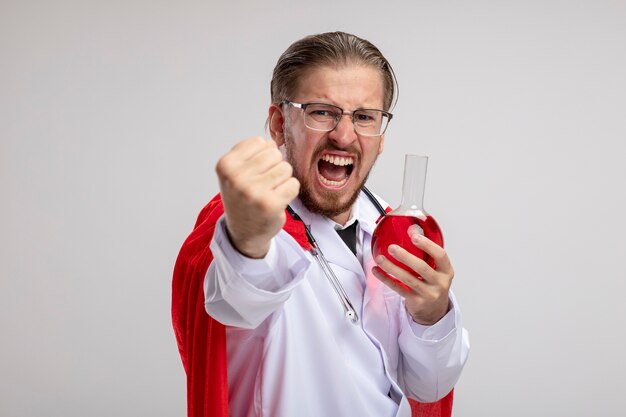 Cara zangado do jovem super-herói vestindo túnica médica com estetoscópio e óculos segurando um copo de química
