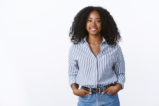 Carismática alegre atraente afro-americana com corte de cabelo encaracolado vestindo uma camisa de mãos dadas, bolsos, confiante, extrovertido, sorrindo, conversando uma conversa agradável, sentindo-se autoconfiante relaxado