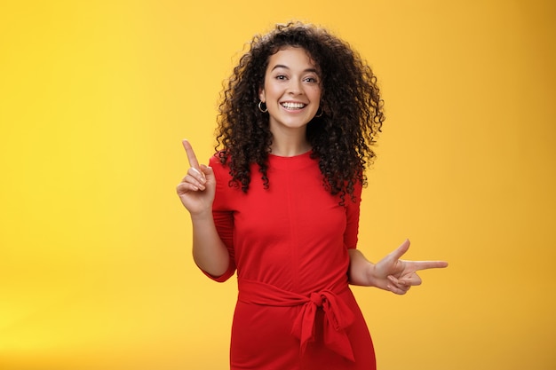 Carismática bonita e feliz jovem fêmea europeia de cabelos cacheados no vestido vermelho, dançando e apontando para cima e para a direita, mostrando de lado como apresentando escolhas, escolhendo, tomando decisões e sorrindo para a câmera.