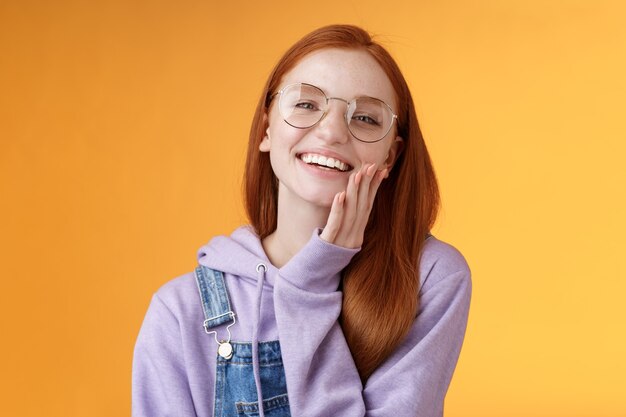 Carismática feliz concurso ruiva sorrindo menina europeia 20 anos óculos com capuz jeans macacão se divertindo aproveitando as férias de verão noite fria rindo alegremente tocar a pele clara pura, fundo laranja.