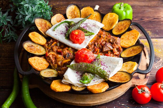 Carne de close-up com berinjela, tomate, batata, pão pita cogumelos e pimenta em um prato redondo em uma mesa de madeira escura horizontal