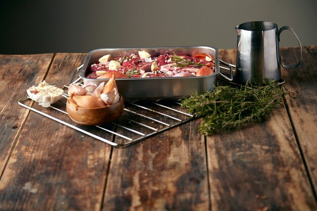 Carne em uma frigideira de aço com temperos ao redor: alho, alecrim, cebola; pronto para cozinhar na mesa de madeira