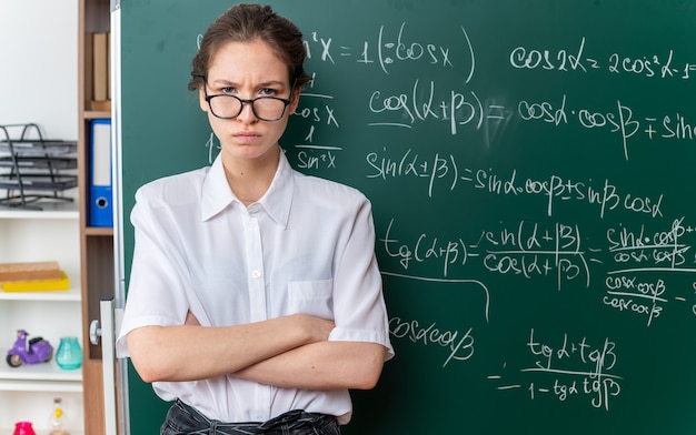 Carrancuda, jovem professora de matemática usando óculos, em pé com a postura fechada em frente ao quadro-negro, olhando para a frente na sala de aula