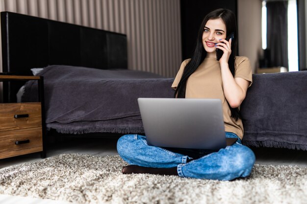 Foto grátis carreira freelance. mulher jovem falando ao telefone usando um laptop, sentada no chão em casa