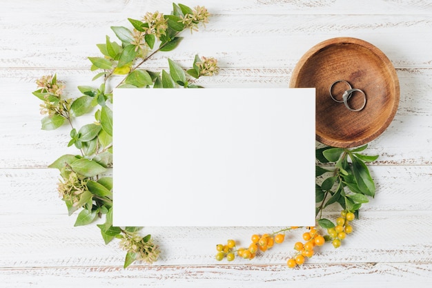 Cartão branco de casamento sobre os anéis; flores e bagas amarelas na mesa de madeira branca