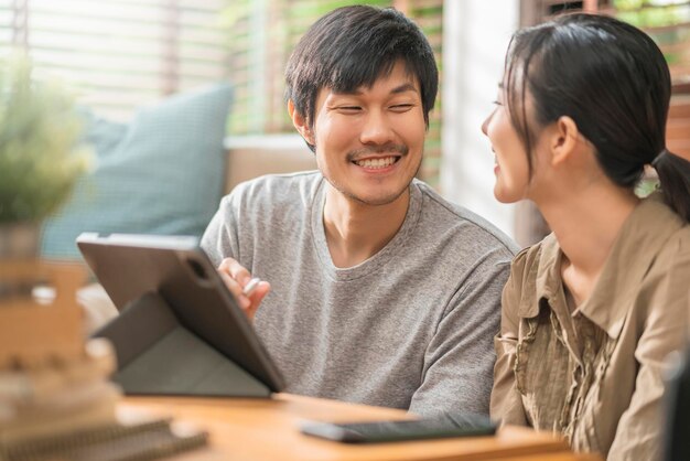 Casa isolar quarentenaasian e amante se casam com casal sentado relaxando usando laptop e tablet trabalhando em casa ou fazendo compras on-line surfando nas mídias sociais com alegre no fundo interior da sala de estar