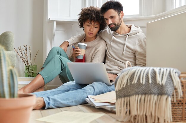 Casal adorável de raça mista se abraçando, sentando no chão e relaxando enquanto assiste ao filme no laptop