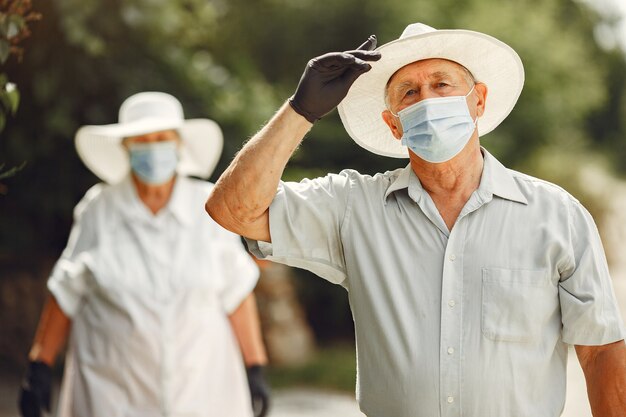 Casal adulto em um jardim de verão. Tema Coromavirus. Pessoas com uma máscara médica. Sênior bonito em uma camisa branca.